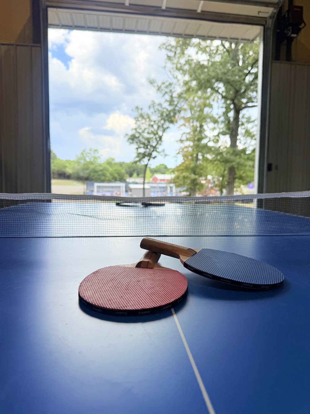 Ping pong table inside the cabin's garage game room, with the garage door rolled up and Pickle Putt Pour's pickleball courts visible outside